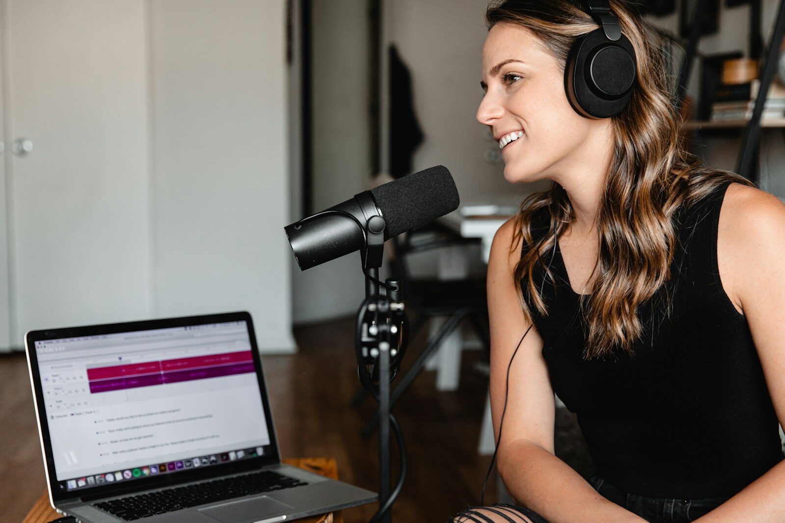 A woman with long hair wearing headphones speaks into a microphone while seated at a desk with a laptop, showcasing a podcasting setup.